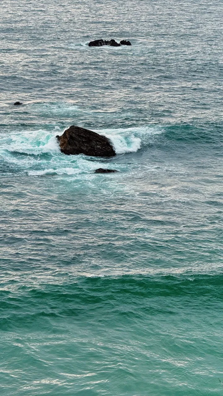 Waves breaking around dark rocks in a teal sea.