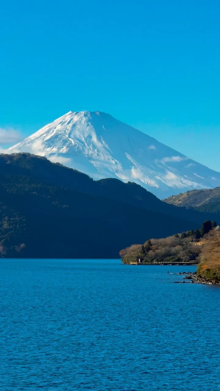 Snow-capped Mount Fuji rising behind a forested ridge and a deep blue lake.