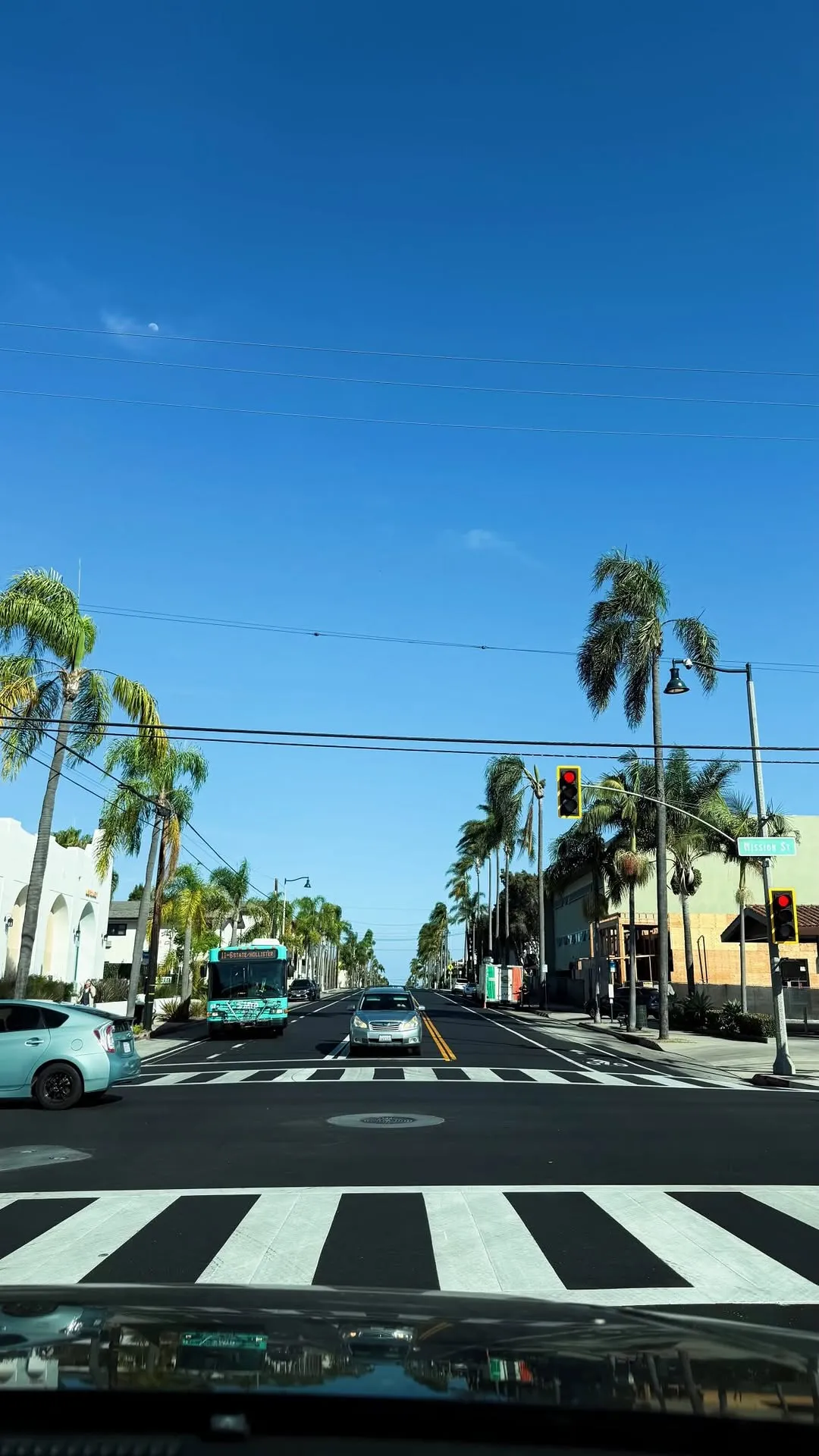 A sunny palm-lined avenue seen from a car at a crosswalk, a local bus and cars ahead under a clear blue sky.