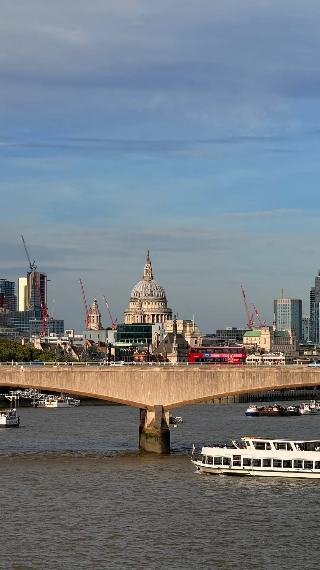 London at golden hour across the Thames, the dome of St Paul's rising behind Waterloo Bridge as a red bus crosses and boats pass below.