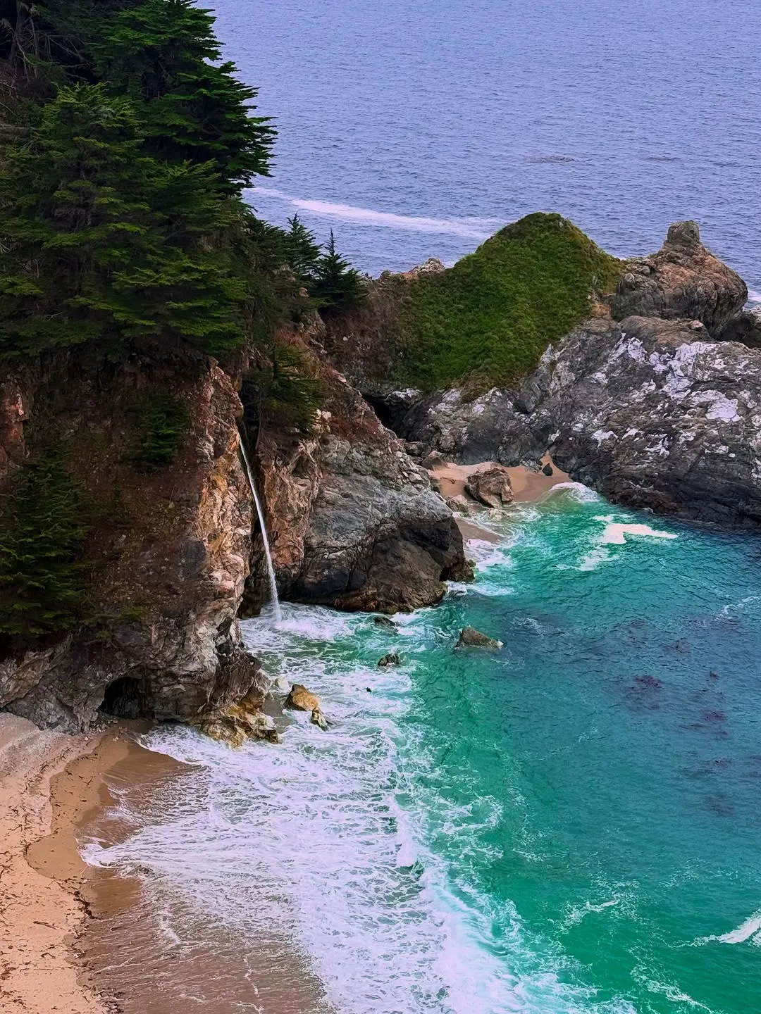 A waterfall tumbling onto a sandy cove of turquoise water, framed by pines on a rocky cliff.